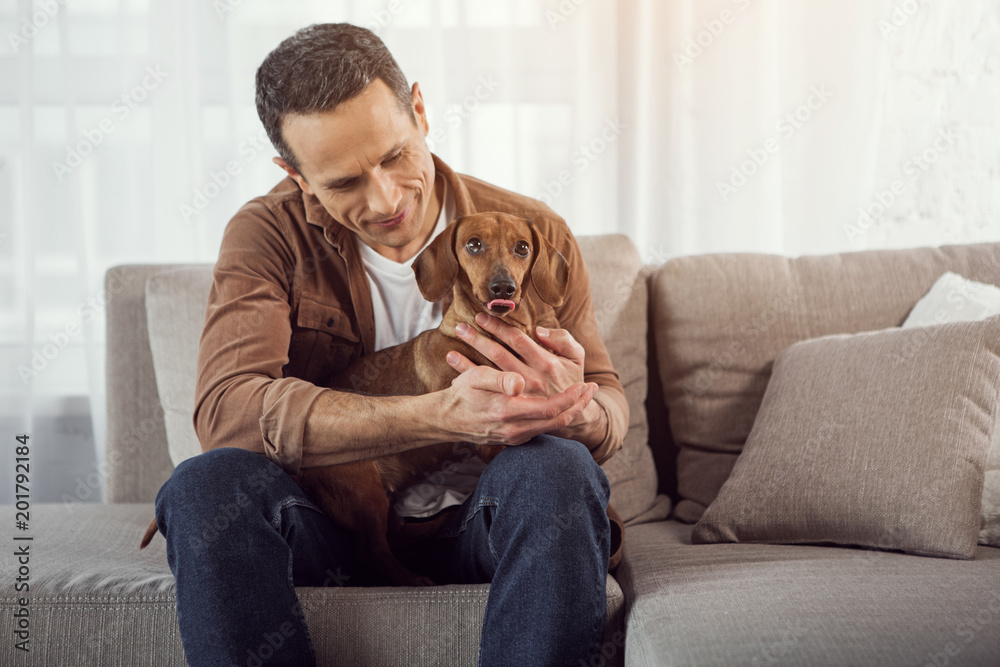 Give me your paw. Portrait of happy man holding his dog and looking at pet with love. He is sitting on sofa and smiling
