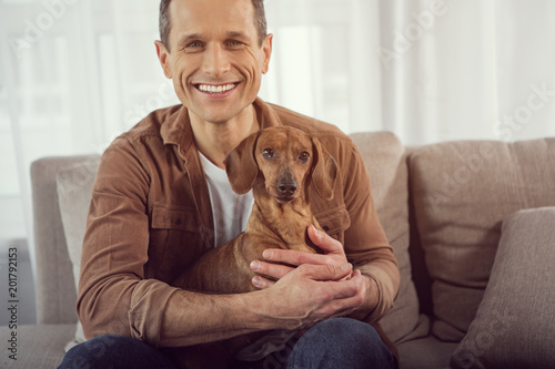 Portrait of excited man keeping puppy in hands and smiling. He is sitting on couch with relaxation