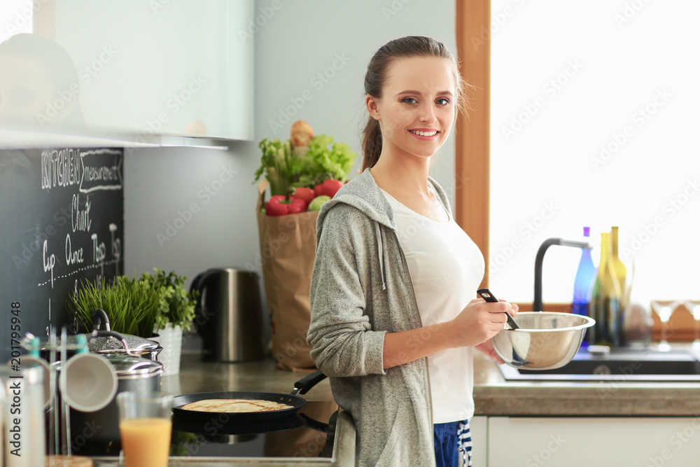 Young woman prepares pancakes in the kitchen while standing near the table. Woman in the kitchen. Cooking at kitchen. Young woman prepares pancakes in the kitchen while standing near the table. Woman in the kitchen. Cooking at kitchen.