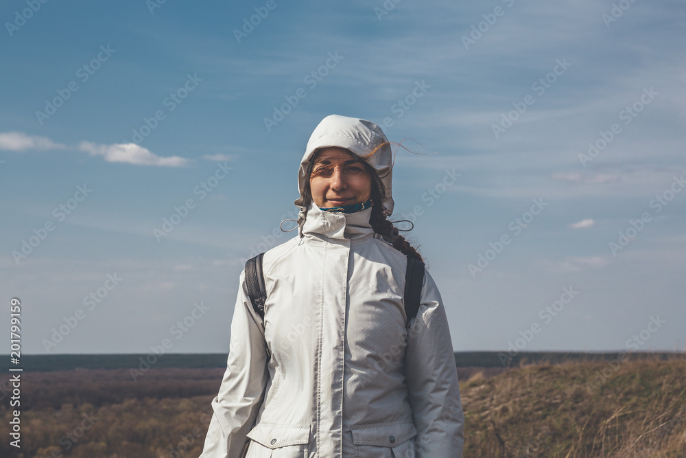 Happy girl traveler in white jacket with hood and backpack smiling and looking at camera on top of windy mountain, travel adventure lifestyle concept