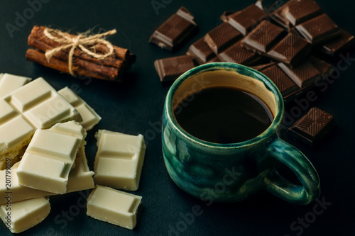 Cup of coffee, broken white and black chocolate bars and stack of cinnamon on black table. Tasty energy calorie food