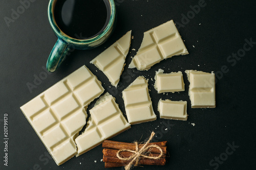 Obraz na plátně Cup of coffee, broken white chocolate bar and stack of cinnamon on black table, top view