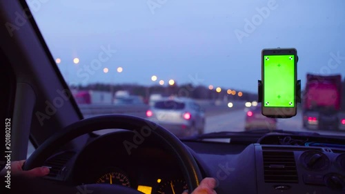 Someone driving a car at night in country. Smartphone with a green screen in the holder on the windshield. Hand of a driver from time to time scrolling the smartphone.