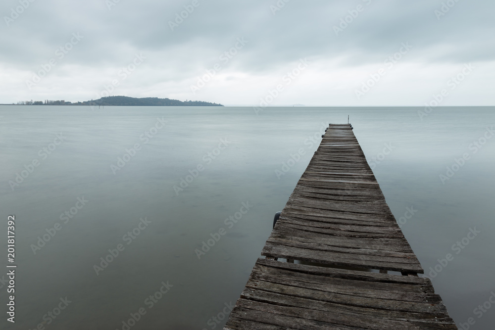 Obraz premium Long exposure first person view of a pier on a lake, with a tree