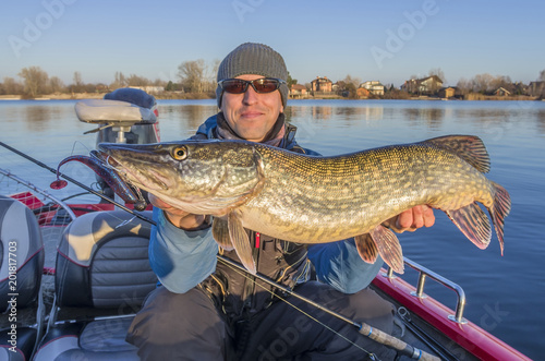 Happy fisherman with big pike fish trophy at the boat with fishing tackles