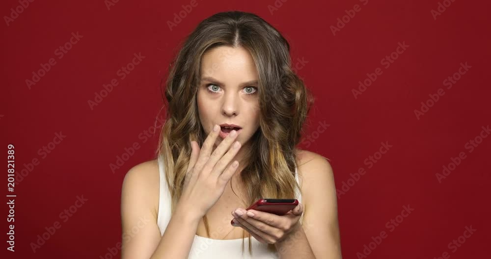Pretty girl looks at mobile phone shocked close up isolated on red background Pretty girl looks at mobile phone shocked close up isolated on red background