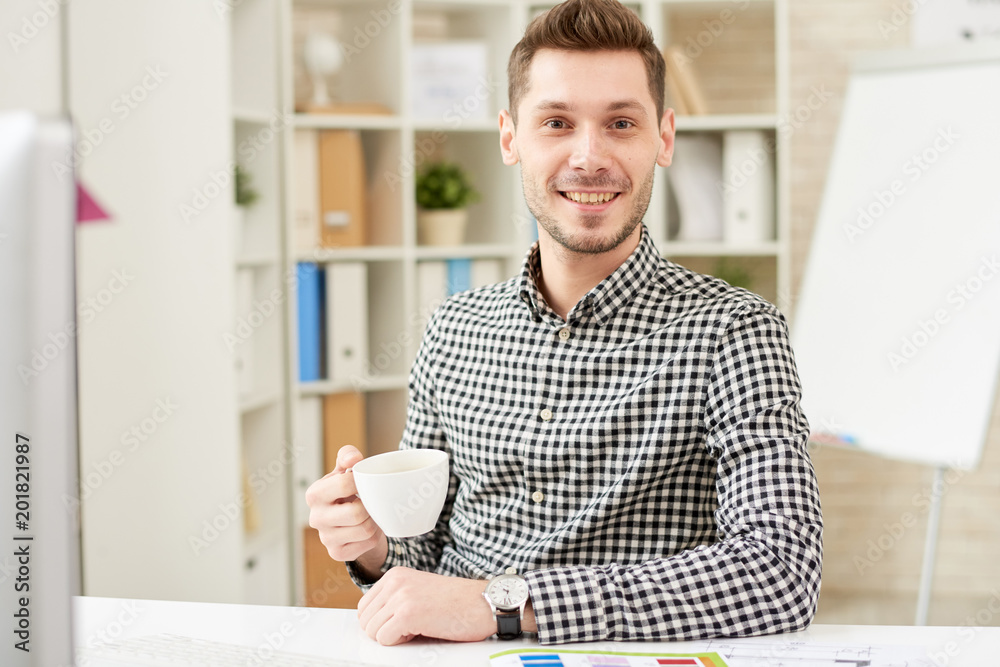 Waist-up portrait of handsome young manager wearing checked shirt looking at camera with wide smile while sitting at office desk and drinking fragrant coffee Waist-up portrait of handsome young manager wearing checked shirt looking at camera with wide smile while sitting at office desk and drinking fragrant coffee