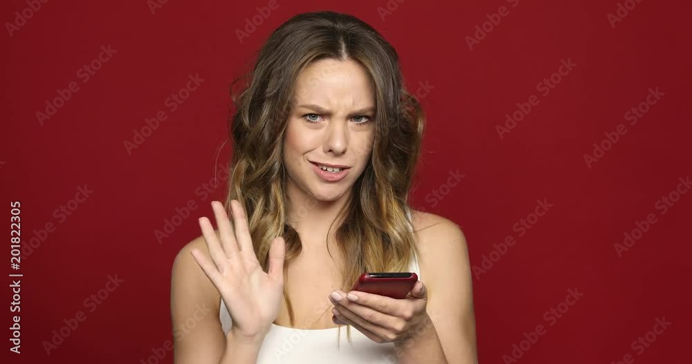 Young woman using her smartphone cellphone surfing the internet see strange news. Girl concerned look in her face looks news on her mobile phone red background Young woman using her smartphone cellphone surfing the internet see strange news. Girl concerned look in her face looks news on her mobile phone red background