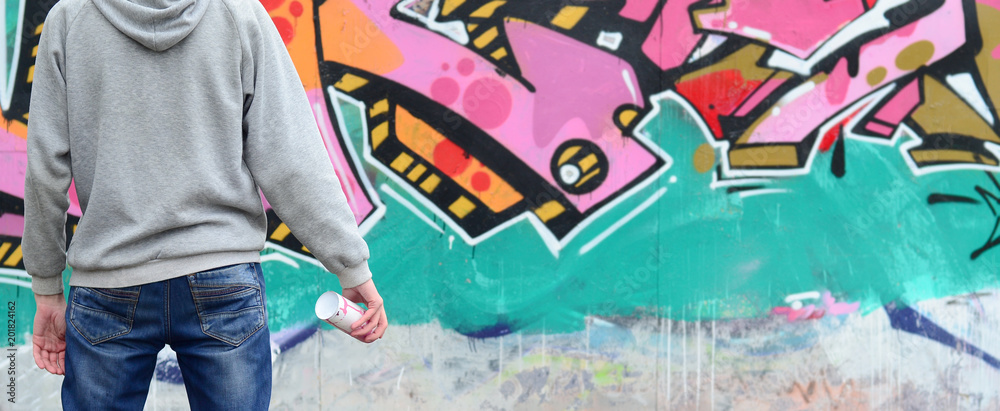 Fototapeta premium Młody graffiti artysta w szarej bluzie z kapturem patrzy na ścianę z jego graffiti w różowe i zielone kolory na ścianie w deszczową pogodę. Koncepcja sztuki ulicy