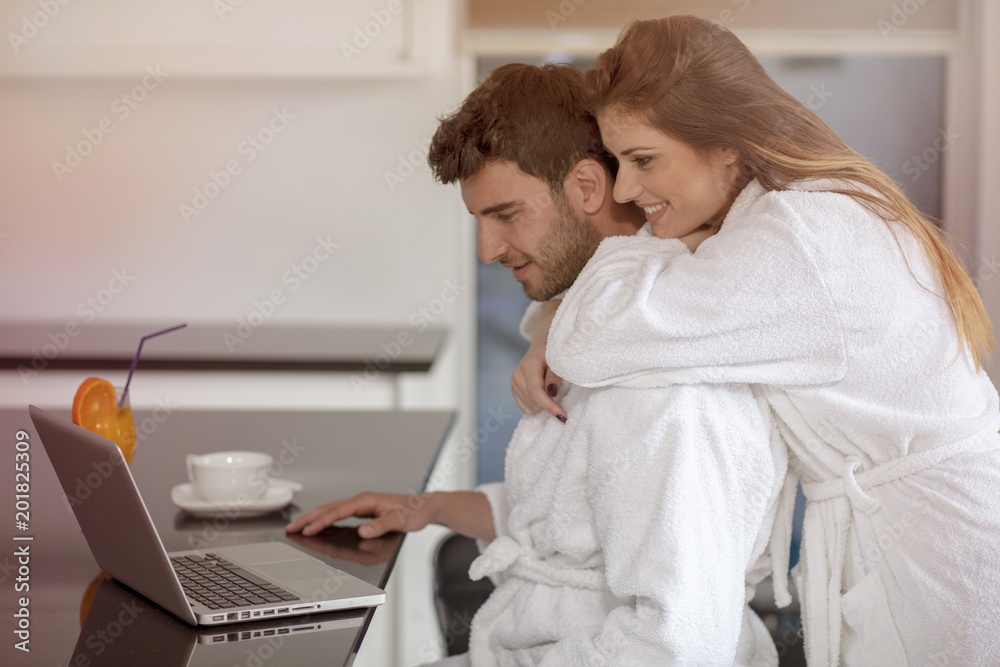 young couple in bathrobe at home having coffee in the kitchen and working on laptop computer young couple in bathrobe at home having coffee in the kitchen and working on laptop computer