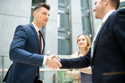 Content handsome young new business employee shaking hand of colleague while getting acquainted with team in office