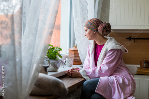 Young adult female cancer patient wearing headscarf and bathrobe sitting in the kitchen with her pet cat, looking out window.