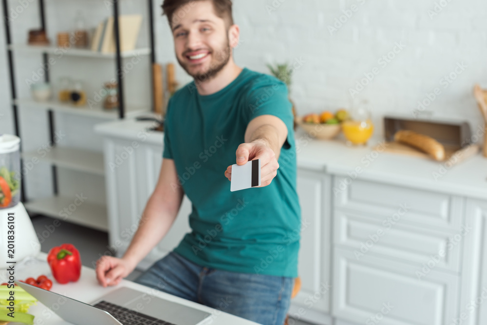 smiling man showing credit card at table with laptop in kitchen at home smiling man showing credit card at table with laptop in kitchen at home
