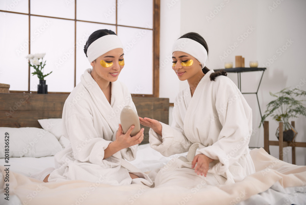 Skin care. Delighted beautiful women holding a bottle with body lotion while looking at it Skin care. Delighted beautiful women holding a bottle with body lotion while looking at it