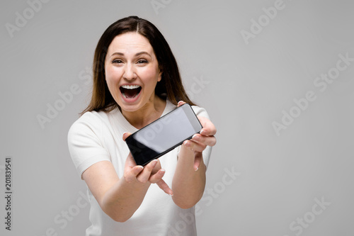 Emotional smiling happy plus size model standing in studio looking in camera offering mobile phone to a client