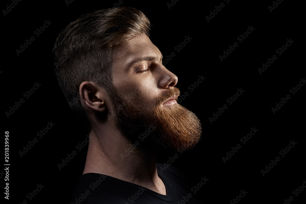Calm peaceful atmosphere. Relaxed attractive male model meditates and tries to relax for minute, keeps eyes shut, dreams about something, isolated over black background