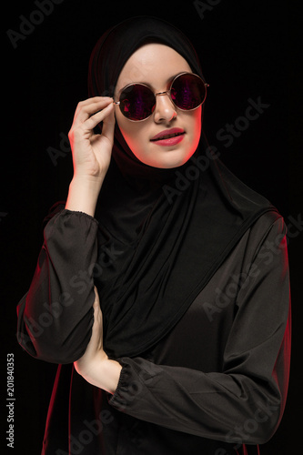 Portrait of beautiful stylish young muslim woman wearing black hijab and sunglasses as modern eastern fashion concept posing on black background