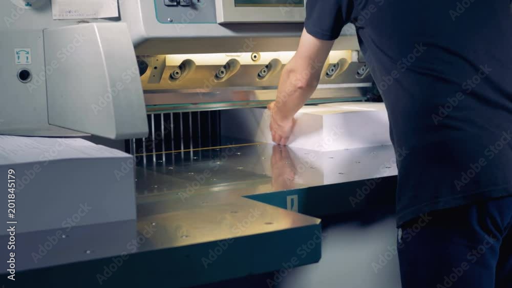 An employee is adjusting two large piles of paper under a cutter which ploughs them in four parts An employee is adjusting two large piles of paper under a cutter which ploughs them in four parts