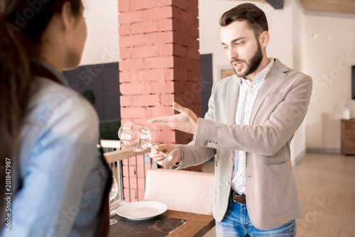 Serious pernickety bearded restaurateur holding examination in modern establishment, strict handsome young restaurant manager making remarks about clearness to waitress photo