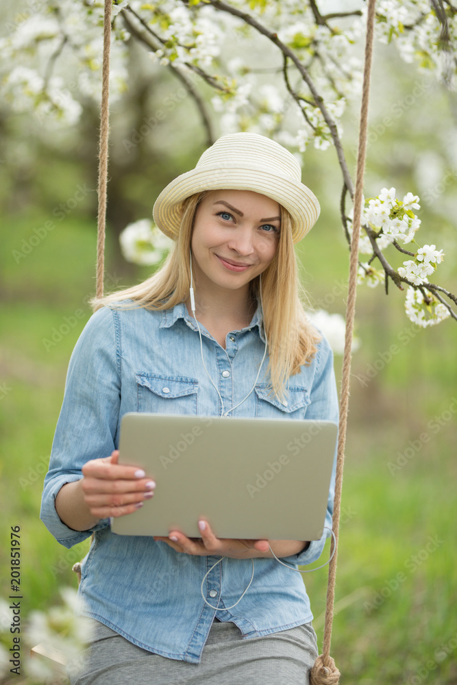 Portrait of a girl in a hat with glasses working on a laptop on a swing, in blooming spring gardens. The concept of freelancing