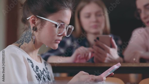 Extravagant student girl with tattoos and wearing glasses is sitting with the phone in the audience, behind her are classmates. A brave modern girl with tattoos surfing the Internet, using smartphone