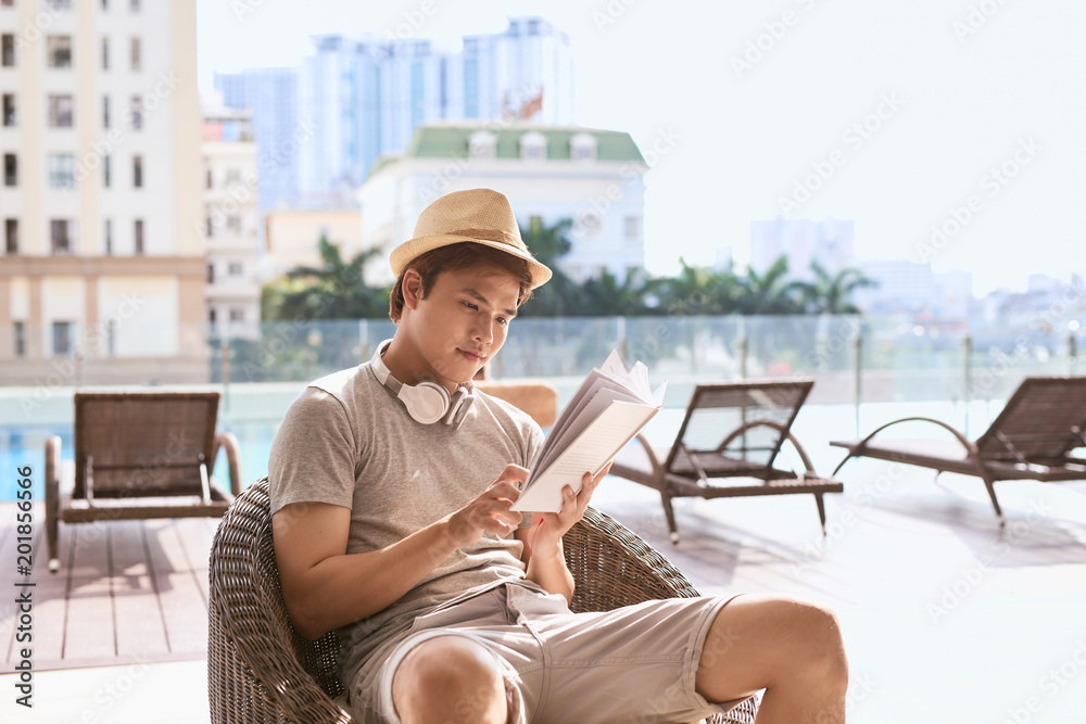 Young asian man reading book by the pool on a sunny summer day