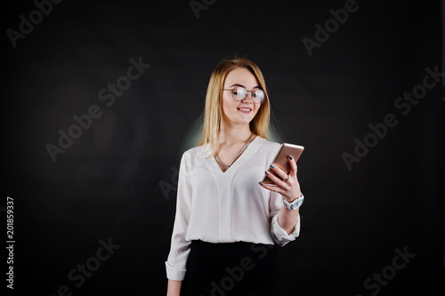 Studio portrait of blonde businesswoman in glasses, white blouse and black skirt looking at mobile phone against dark background. Successful woman and stylish girl concept.