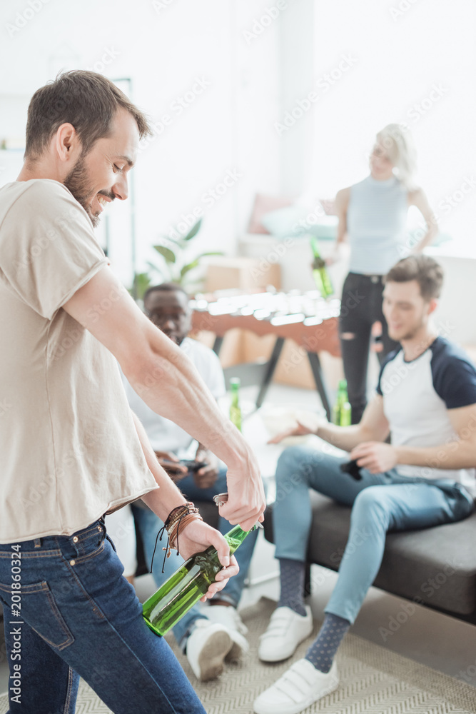© LIGHTFIELD STUDIOS - side view of smiling young man opening beer bottle by corkscrew and friends sitting near © LIGHTFIELD STUDIOS - side view of smiling young man opening beer bottle by corkscrew and friends sitting near