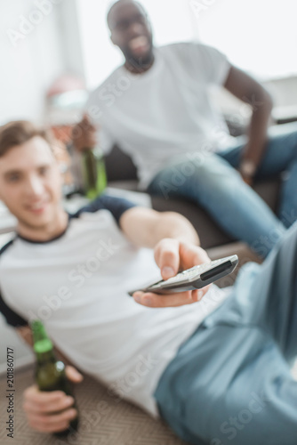 closeup shot of remote in hand of man sitting with bottle of beer near friend