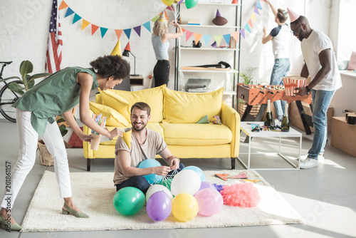 young african american woman proposing party hat to smiling man on floor with balloons and woman with friend