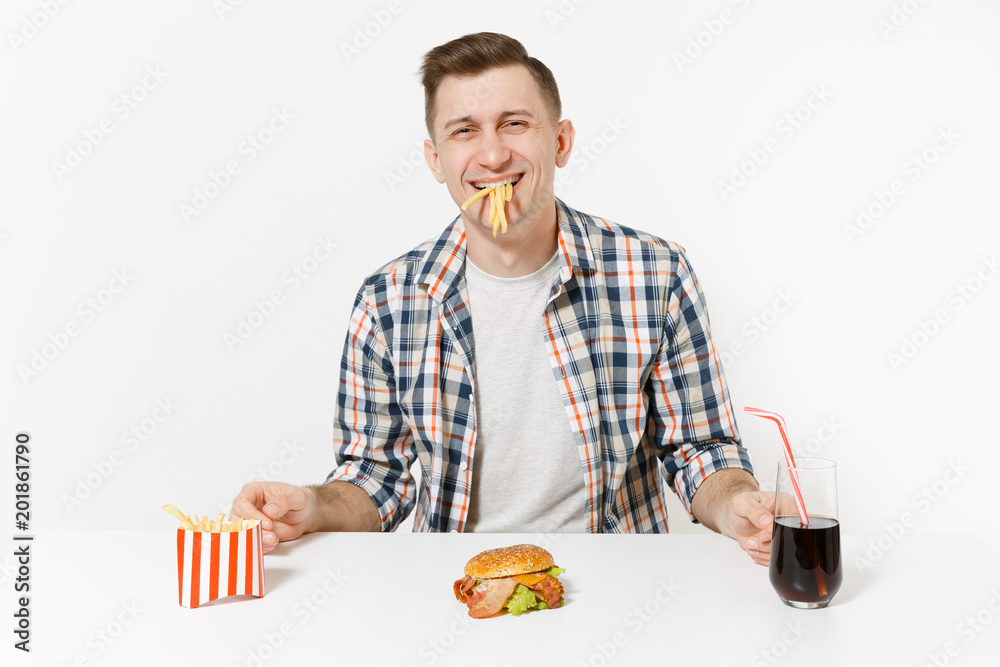 Fun hungry young man eating french fries, sitting at table with burger, cola in glass isolated on white background. Proper nutrition or American classic fast food. Advertising area with copy space. Fun hungry young man eating french fries, sitting at table with burger, cola in glass isolated on white background. Proper nutrition or American classic fast food. Advertising area with copy space.