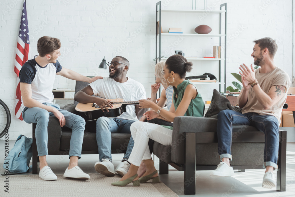 group of multiethnic smiling young people playing guitar in living room group of multiethnic smiling young people playing guitar in living room