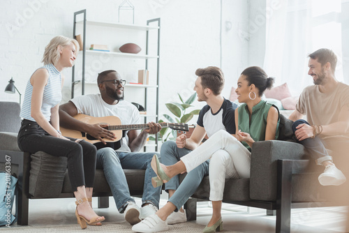 multicultural group of young people sitting in living room and playing acoustic guitar