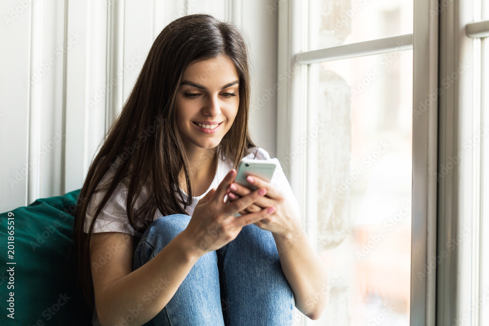 Beautiful smiling brunette girl wears white t-shirt using smartphone sitting on the windwsill Beautiful smiling brunette girl wears white t-shirt using smartphone sitting on the windwsill