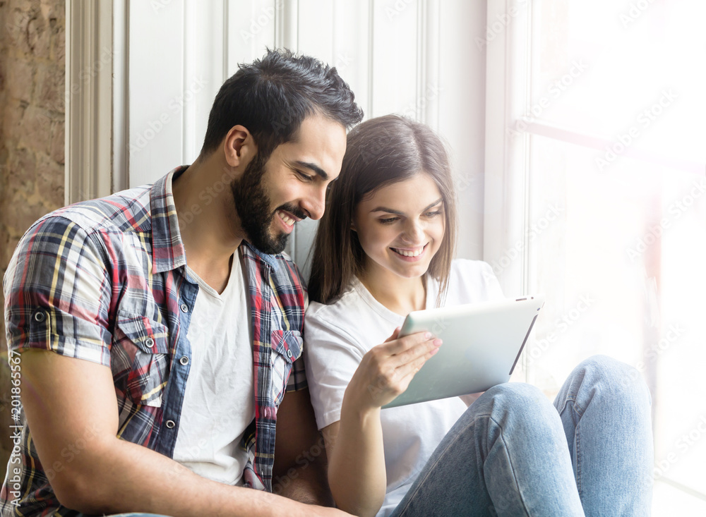 Bearded handsome man and brunette girl sitting on the wide windowsill using tablet computer Bearded handsome man and brunette girl sitting on the wide windowsill using tablet computer