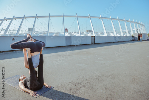 Young athletic couple practicing acroyoga at stadium urban style.Balancing in pair. Fit active pair yoga time.Sporty handsome man supporting slim beautiful brunette woman.Training time.Asana 
