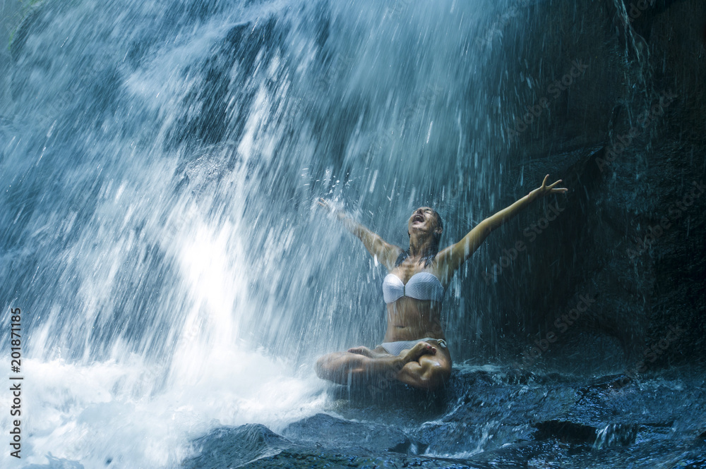 Obraz premium attractive woman sitting at rock in yoga pose for spiritual relaxation serenity and meditation at stunning beautiful waterfall and rain forest in Bali