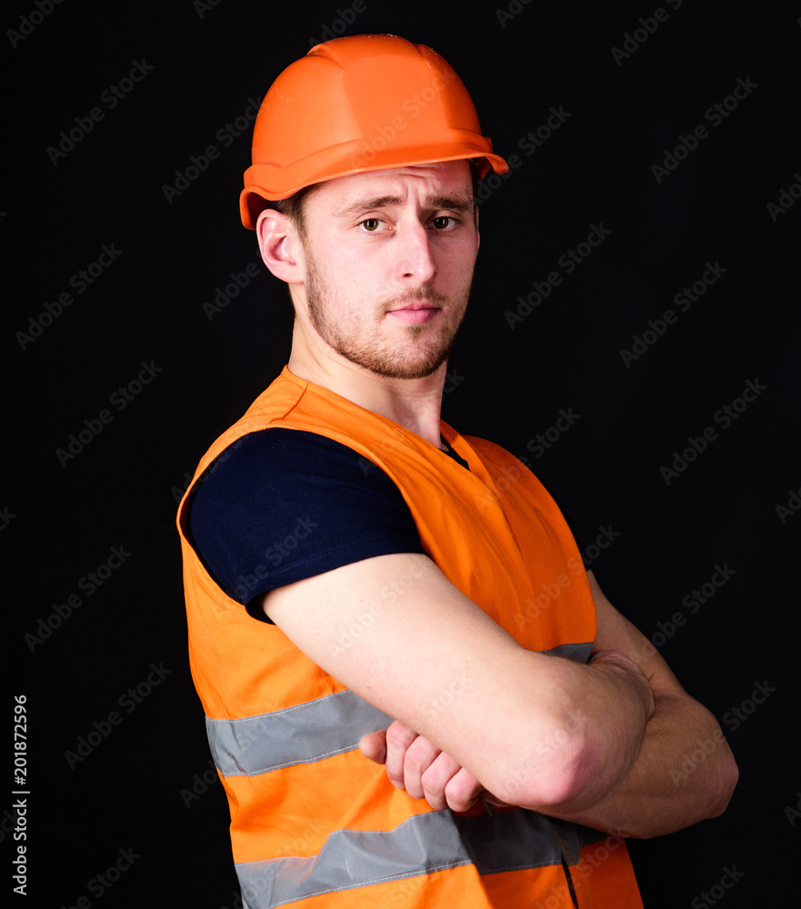 Strong builder concept. Builder in helmet posing confidently. Man in helmet, hard hat hold arms crossed on chest, black background. Worker, contractor, builder on serious face with muscular biceps. Strong builder concept. Builder in helmet posing confidently. Man in helmet, hard hat hold arms crossed on chest, black background. Worker, contractor, builder on serious face with muscular biceps.