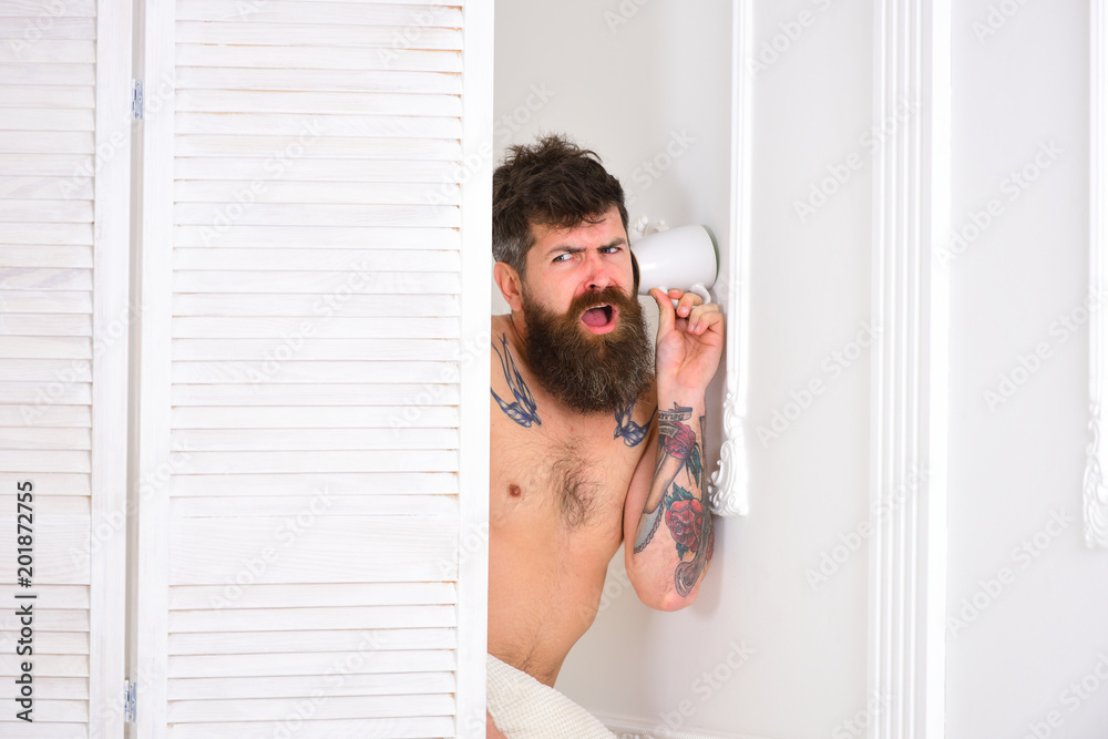 Lovers and cheating concept. Man with beard and mustache eavesdrops using mug near wall. Man in white interior spying, eavesdropping. Hipster naked on stunned face secretly listen conversation. Lovers and cheating concept. Man with beard and mustache eavesdrops using mug near wall. Man in white interior spying, eavesdropping. Hipster naked on stunned face secretly listen conversation.