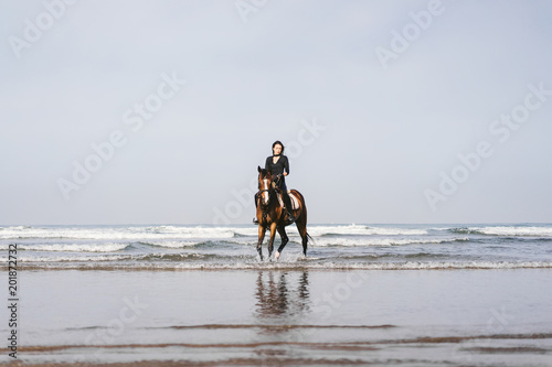 distant view of woman riding horse on sandy beach with ocean behind