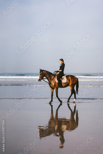 young female equestrian riding horse on sandy beach near ocean