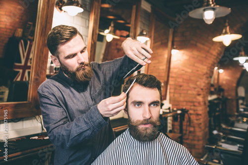 A picture of two bearded guys are in a barbershop. The hairdresser is cutting the hair of his customer using scissors and small hair brush.