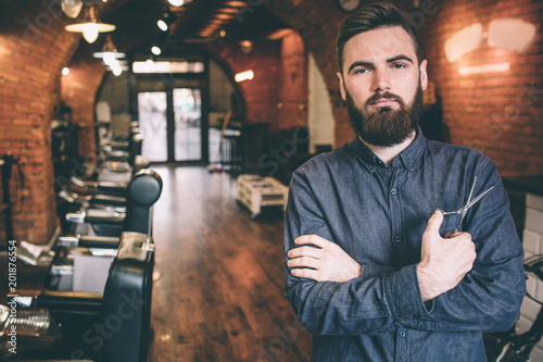 Beautiful picture of handsome guy is standing in a barbershop and keepinh his hands crossed. He is holding a pair of scissors in the right hand.