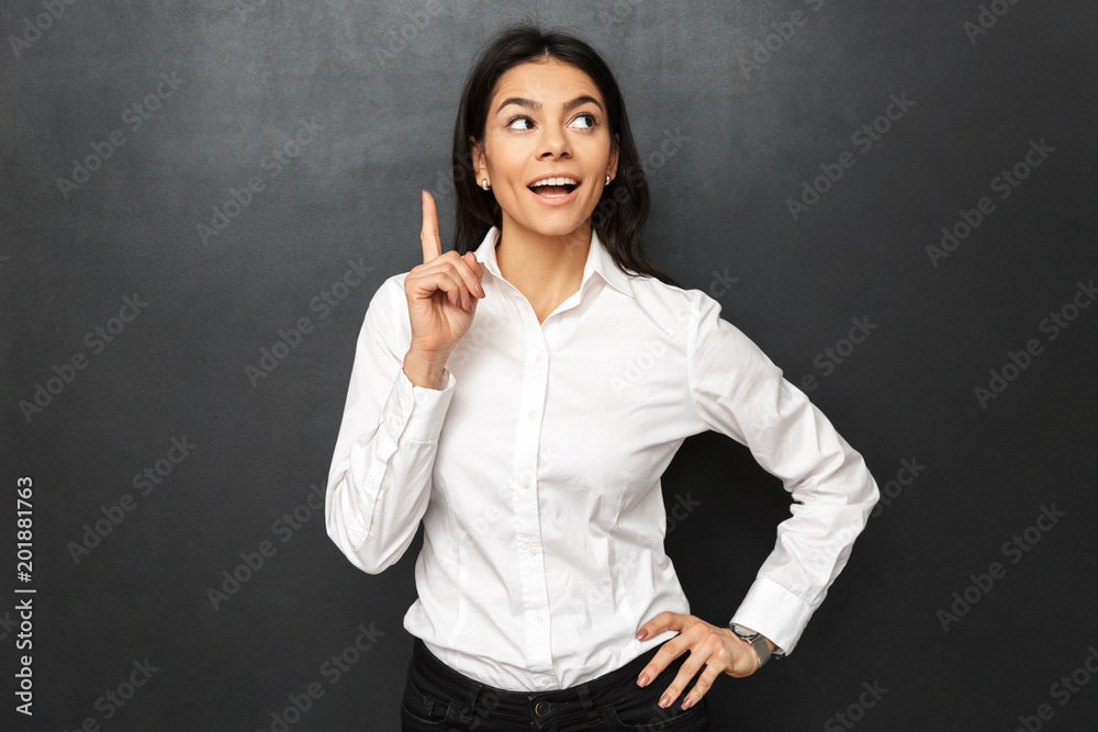 Portrait of business young woman wearing white shirt showing index finger up like having idea, isolated over dark gray background Portrait of business young woman wearing white shirt showing index finger up like having idea, isolated over dark gray background