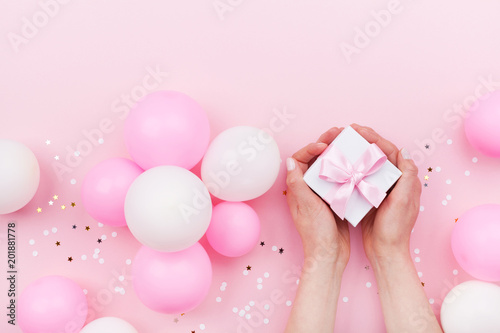 Womans hands holding gift or present box on pink pastel table decorated balloons and confetti. Flat lay style. © juliasudnitskaya