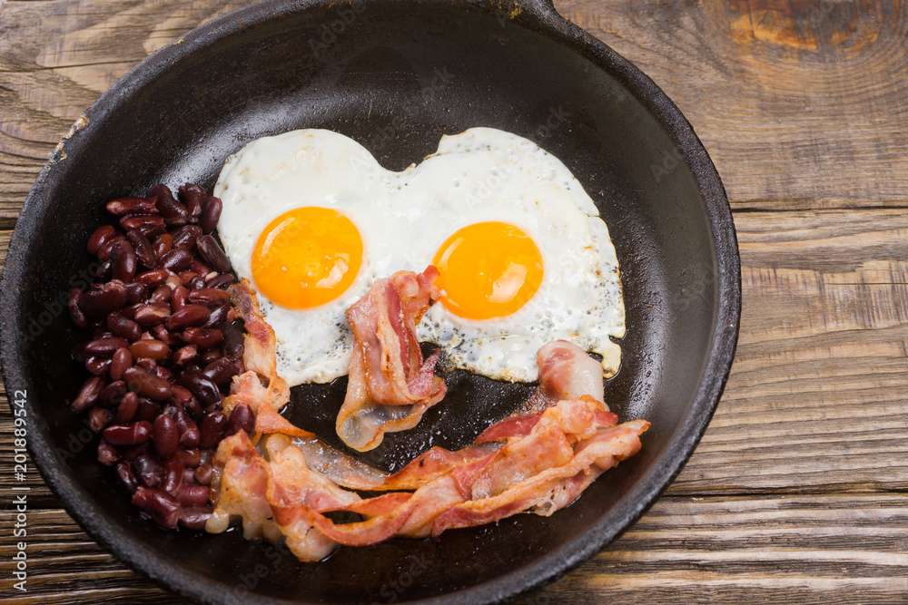 Bacon,Egg and bean. Salted egg and sprinkled with black pepper. English breakfast. Grilled bacon, two eggs and beans in pan on wooden table Bacon,Egg and bean. Salted egg and sprinkled with black pepper. English breakfast. Grilled bacon, two eggs and beans in pan on wooden table