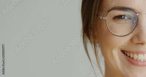 Close up of the half female facen in glasses with long fair hair looking in the camera and then smiling on the white wall background. Indoor photo