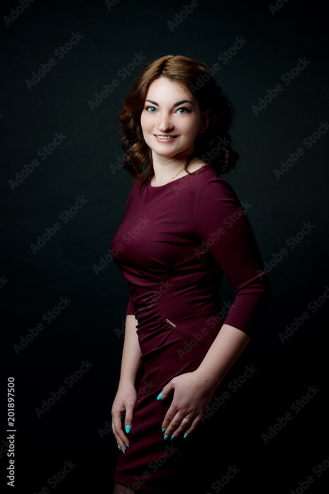 A brunette woman with makeup and hairdo in a claret dress smiles against a black background. A brunette woman with makeup and hairdo in a claret dress smiles against a black background.