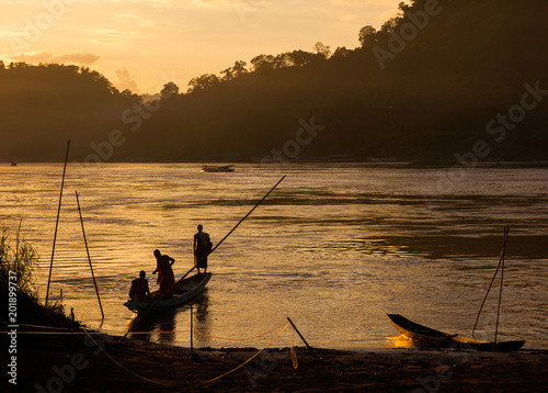 Shadowy sunset view of Mekong river and traditional boat with buddhist monks. River bank covered with jungle background. Luang Prabang, Laos.