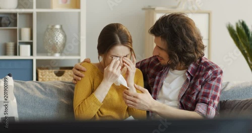 Young attractive couple sitting on the sofa, watching a melodrama movie and man calming down a woman as she crying. Indoor photo